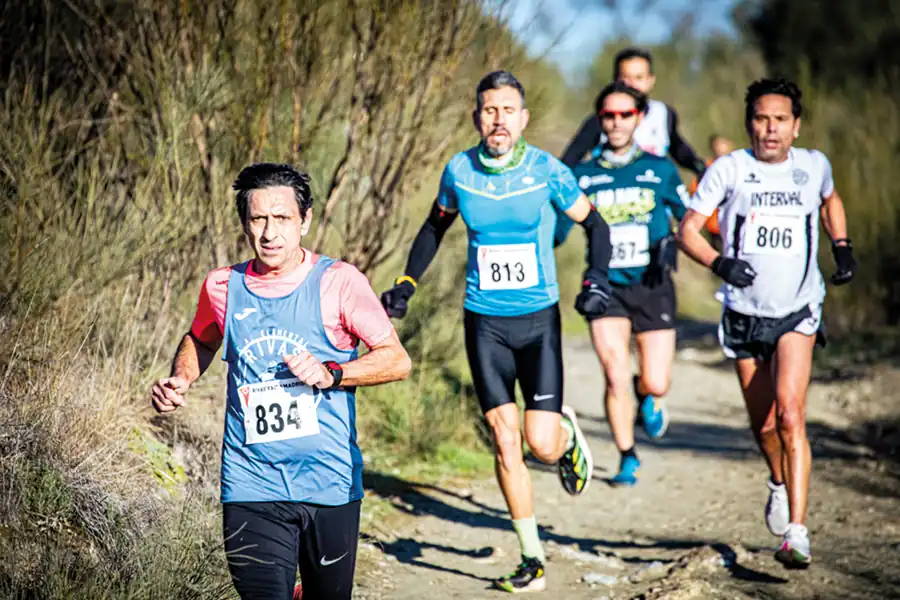 Cuatro hombres compiten en el Cross de Rivas, corriendo por un sendero de tierra bordeado de vegetación. Ataviados con dorsales numerados y ropa de atletismo, encarnan el espíritu del atletismo popular bajo la brillante luz del sol.