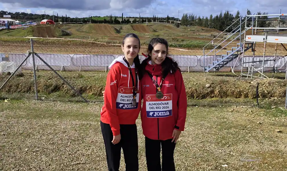 Dos jóvenes atletas con chaquetas rojas y pantalones negros posan uno al lado del otro en un cross estatal en Ciudad de Rivas, posando en un campo de hierba con dorsales, barreras metálicas y escaleras visibles al fondo.