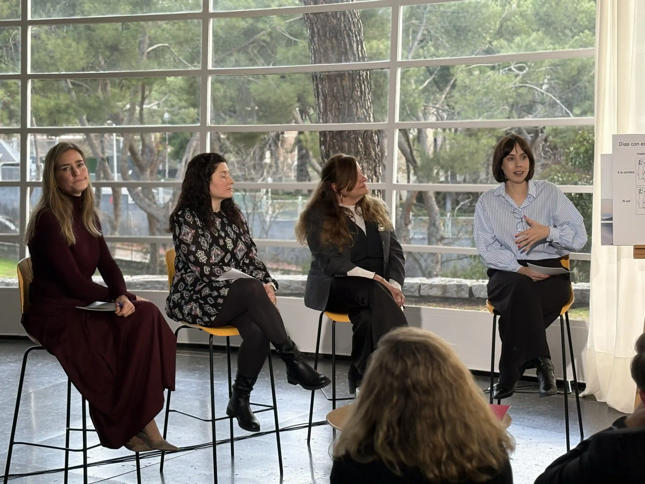 Cuatro mujeres sentadas en taburetes frente a grandes ventanales durante una mesa redonda sobre ciencia climática. Una mujer a la derecha habla, mientras las otras escuchan atentamente. El público está sentado en primer plano. Cuatro mujeres sentadas en taburetes frente a grandes ventanales durante una mesa redonda sobre ciencia climática. Una mujer a la derecha habla, mientras las otras escuchan atentamente. El público está sentado en primer plano.