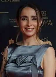 Una mujer con el pelo largo y castaño lleva un vestido plateado sin mangas y sonríe a la cámara mientras está de pie delante de un fondo oscuro con logotipos y texto de Zahara y Fiestas de Mayo Rivas.