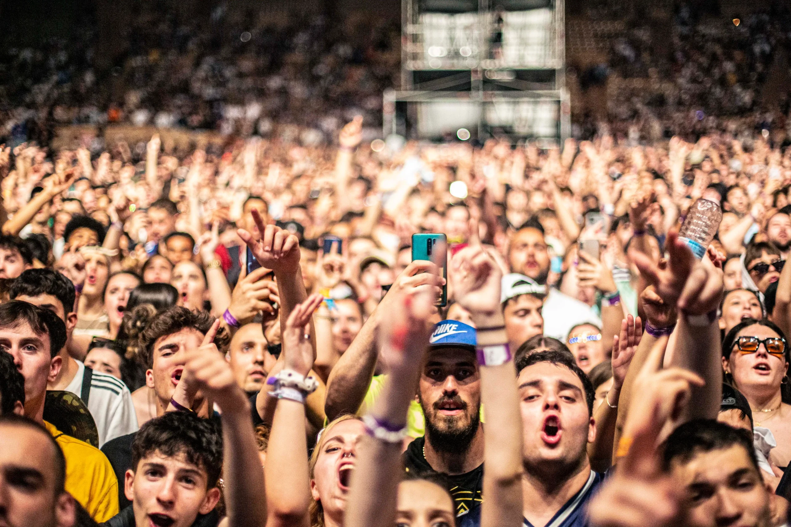 Una gran multitud en las Fiestas de Mayo de Rivas aplaude y levanta los brazos cuando Zahara y Ojete Calor iluminan el escenario, y muchos capturan el momento con sus teléfonos. Al fondo se ven más espectadores emocionados y el equipo del escenario.