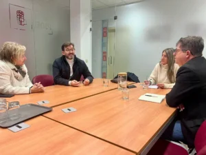 Cuatro personas se sientan alrededor de una mesa de conferencias en la sala de reuniones de una oficina, discutiendo ideas para apoyar el tejido empresarial local. Sobre la mesa hay vasos de agua, cuadernos y bol&iacute;grafos. Al fondo, paredes de cristal y una puerta.
