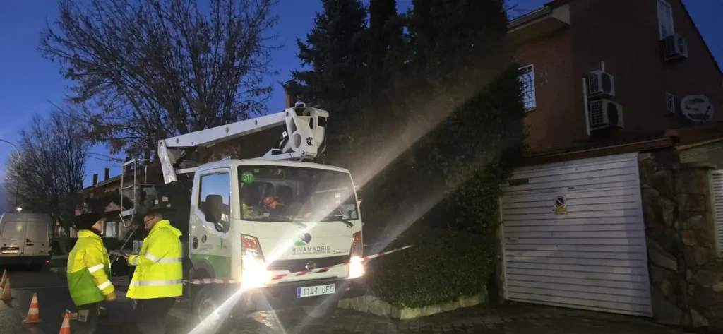 Un camión de servicio público con una plataforma de trabajo elevada está aparcado junto a la acera al anochecer. Dos trabajadores con chalecos fluorescentes están cerca, y al fondo se ve una casa con garaje, donde podrían estar guardadas las herramientas del Borrador automático.