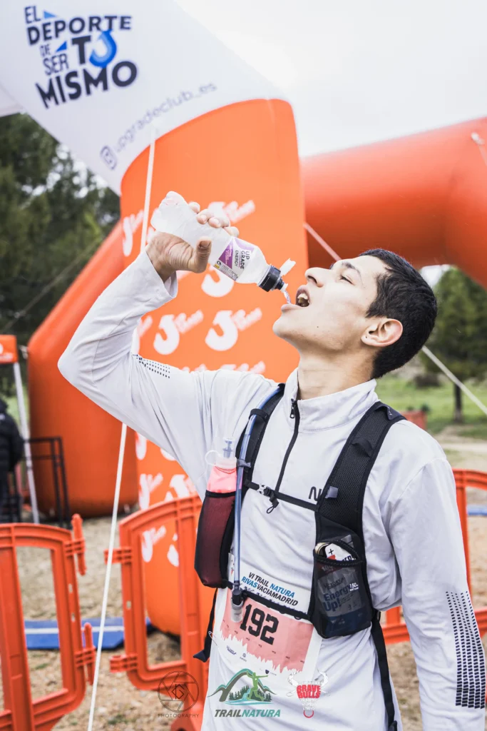 Un corredor con el dorsal 192 bebe de una botella de agua cerca de un arco hinchable naranja en el Rivas Trail Natura 2026. El participante se une a otros corredores con una chaqueta blanca y un chaleco de hidratación en este evento deportivo al aire libre.