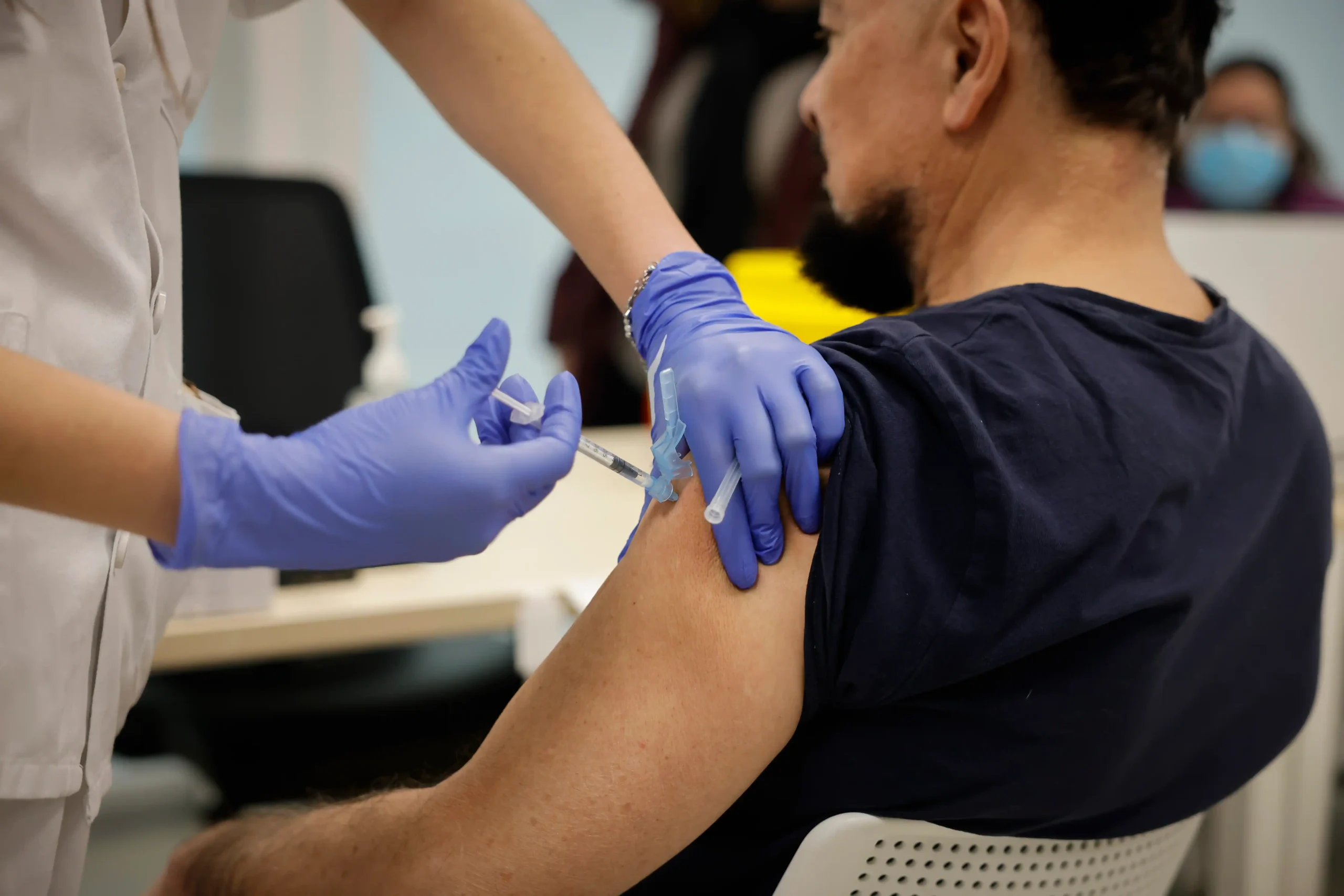 Una persona con camisa oscura recibe una inyección en la parte superior del brazo de un trabajador sanitario que lleva guantes. El entorno parece ser una clínica o un centro médico. Una persona con camisa oscura recibe una inyección en la parte superior del brazo de un trabajador sanitario que lleva guantes. El entorno parece ser una clínica o un centro médico.