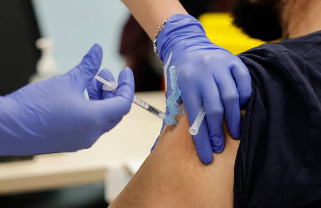 Una persona con guantes azules administra una inyección de vacuna en la parte superior del brazo de otra persona, utilizando una jeringuilla y una aguja, mientras sostiene cerca un borrador automático.