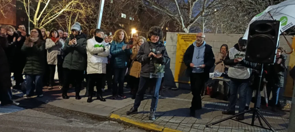 Un grupo de personas de pie en una acera de la ciudad por la noche en Rivas, algunos aplaudiendo y otros sosteniendo papeles sobre la sanidad pública. Se ven árboles y edificios desprovistos de hojas, con un altavoz y una mesa de personas a la derecha, lo que pone de relieve la urgencia de su reunión.