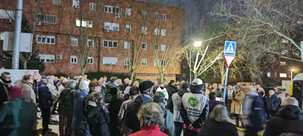 Una multitud se reúne al aire libre frente a un edificio de apartamentos de ladrillo por la noche en Rivas. Algunos llevan abrigos y sombreros, lo que subraya la urgencia del momento. Los árboles desnudos y las señales de tráfico destacan bajo el resplandor de las farolas.