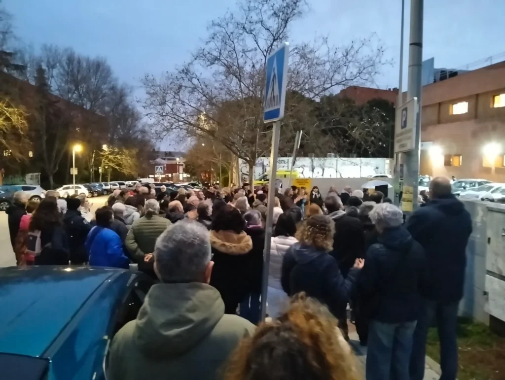 Una multitud de personas permanece de pie en una acera y una calle al atardecer, de espaldas a la cámara, como si esperaran una urgencia. Al fondo se ven árboles deshojados, coches aparcados y edificios bajo un cielo nublado.