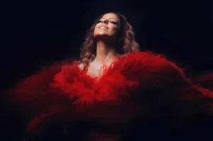 Una persona con el pelo largo y ondulado sonríe mientras lleva o está rodeada por una espectacular prenda de plumas de color rojo brillante sobre un fondo oscuro.