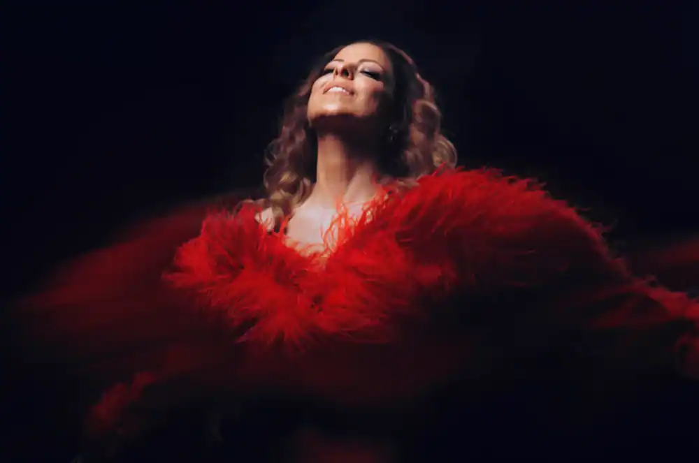 Una persona con el pelo largo y ondulado sonríe mientras lleva o está rodeada por una espectacular prenda de plumas de color rojo brillante sobre un fondo oscuro.