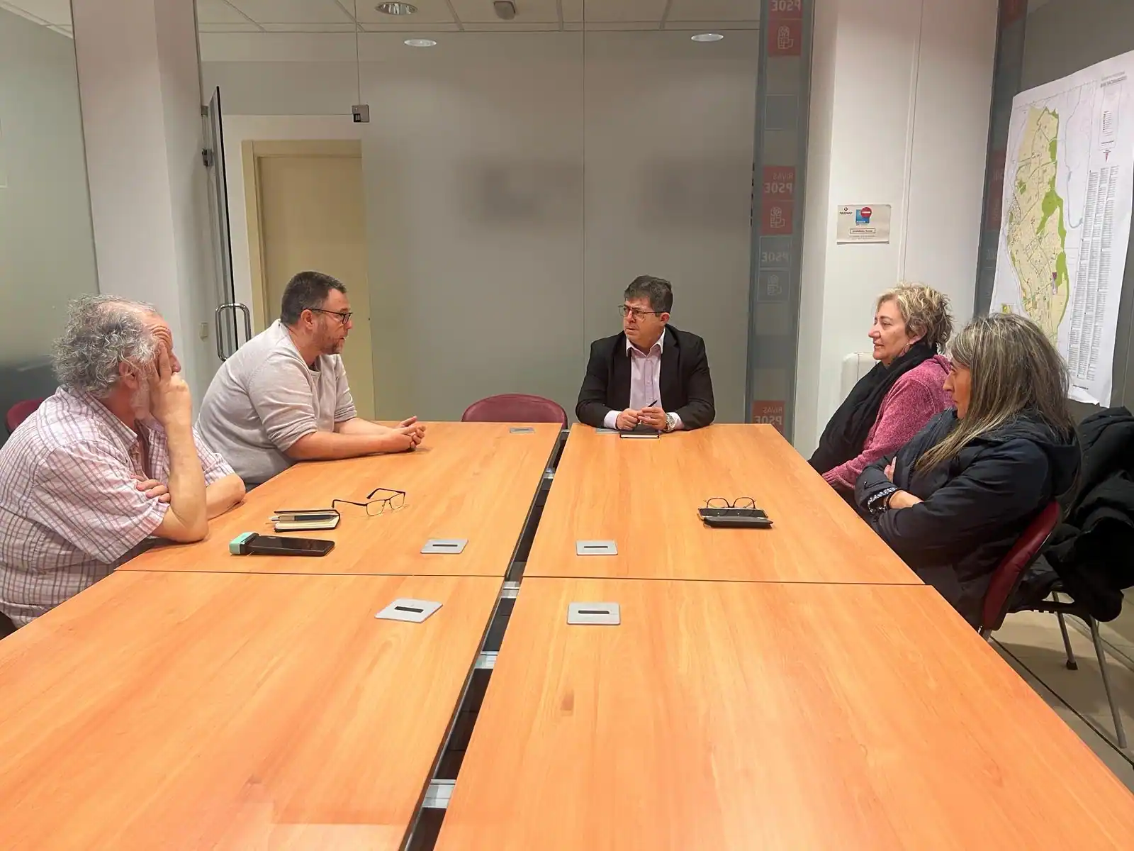 Cinco personas se sientan alrededor de una mesa de reuniones rectangular de madera en una oficina, enfrascadas en una conversación. Sobre la mesa hay papeles, gafas, un teléfono y un borrador automático. Un mapa cuelga de la pared de la derecha. Cinco personas se sientan alrededor de una mesa de reuniones rectangular de madera en una oficina, enfrascadas en una conversación. Sobre la mesa hay papeles, gafas, un teléfono y un borrador automático. Un mapa cuelga de la pared de la derecha.