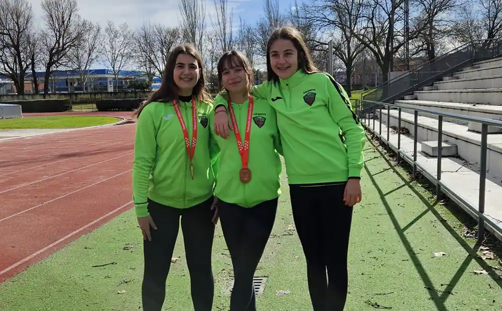Tres jóvenes atletas, entre ellas Lidia Bravo y Heba Melero, con chaquetas verdes a juego, de pie en una pista de atletismo. Dos de ellas llevan medallas al cuello y sonríen para la foto, con las gradas visibles al fondo.