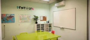 Un aula de la academia Pentalfa tiene paredes verde pálido, una mesa verde lima con sillas, una pizarra blanca, una tabla periódica, un mapamundi, una planta, estanterías con material y un tablón de anuncios, ideal para el aprendizaje escolar. "PENTALFA" está pintado en la pared.