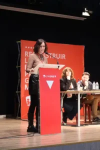 Una mujer de pie en un podio rojo de IU Madrid, presenta la hoja de ruta 2027 a un público. Dos personas se sientan en una mesa detrás de ella, con un telón de fondo rojo y texto de fondo, lo que sugiere un evento o una conferencia en interiores.