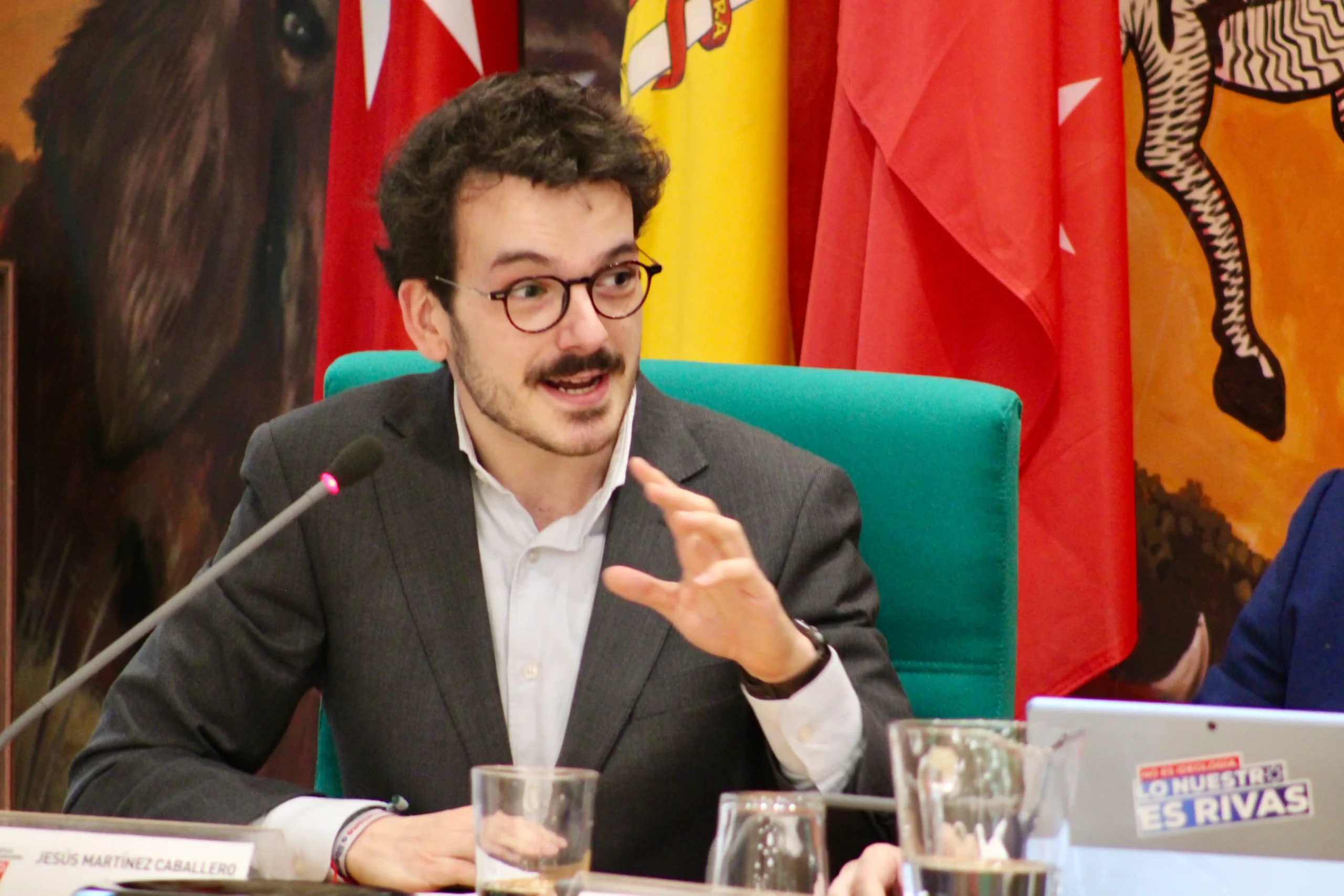 Un hombre con gafas y bigote, identificado como el portavoz adjunto del PP Jesús Martínez Caballero, habla por un micrófono sentado en una mesa con una silla verde. En el fondo se ven banderas y su placa identificativa.