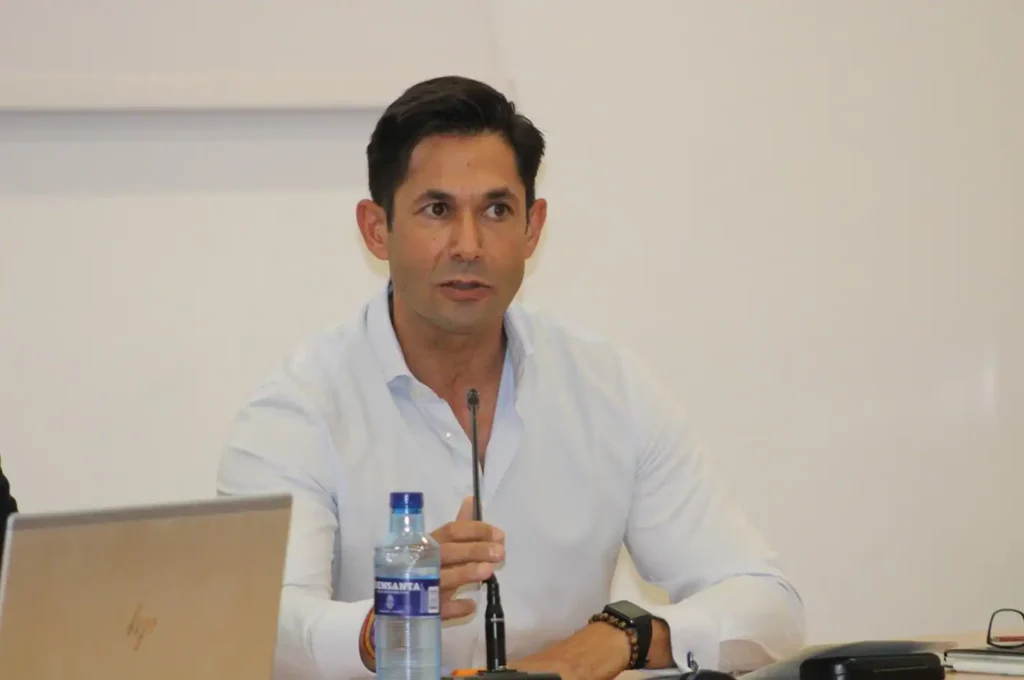 Jesús Martínez Caballero, portavoz adjunto PP, se sienta a una mesa hablando frente a un micrófono. Sobre la mesa hay un portátil y una botella de agua. El fondo es completamente blanco y sencillo.