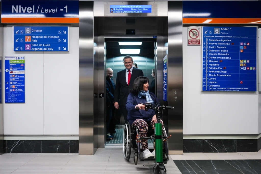 Una mujer en silla de ruedas sale de un ascensor accesible, seguida de dos hombres en traje de negocios en la estación de Metro de Avenida América, con señalización bilingüe en español e inglés.