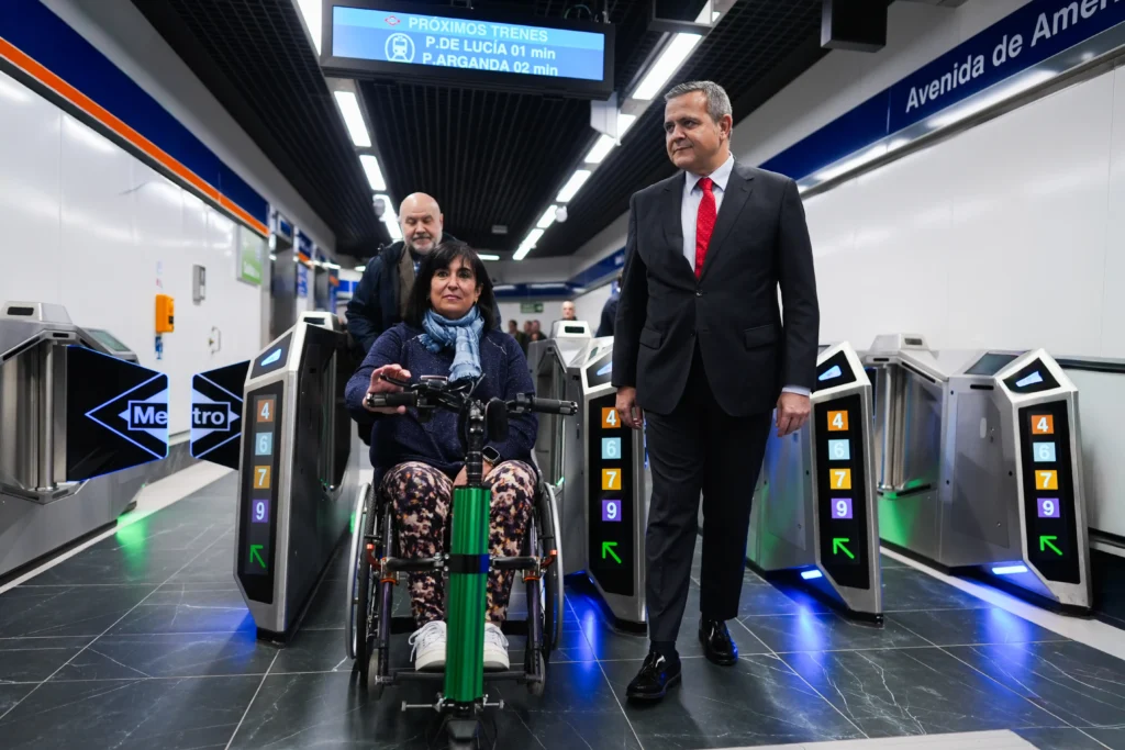 Una mujer en silla de ruedas, un hombre trajeado y otro hombre caminan juntos por la estación de Metro de Avenida América, una estación accesible, bien iluminada, con señalización visible y marcadores numerados junto a los torniquetes.