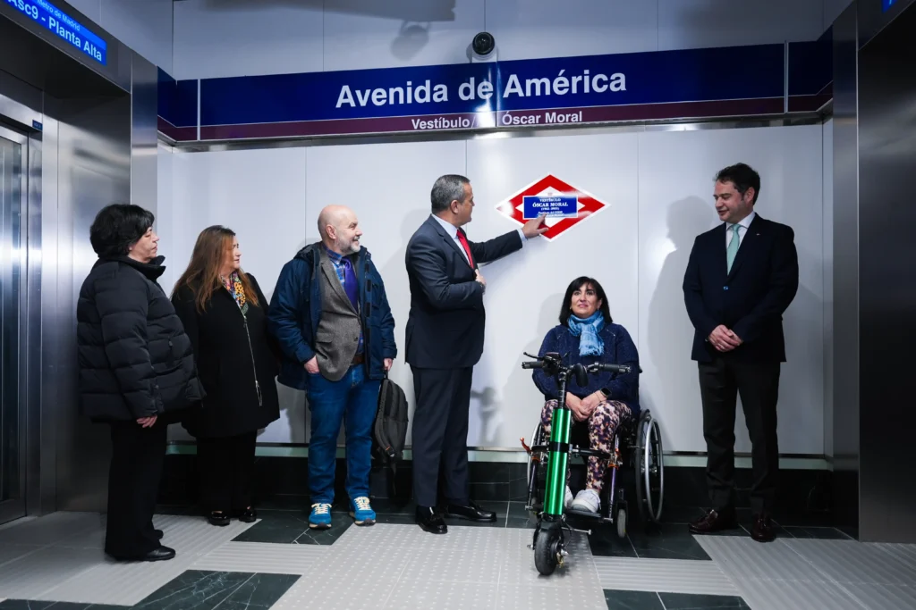 Seis personas permanecen de pie en la estación de Metro de Avenida América bajo una señal. Un hombre señala un cartel, mientras una mujer en silla de ruedas con un accesorio verde se sienta cerca del centro. El grupo parece estar en un evento accesible.