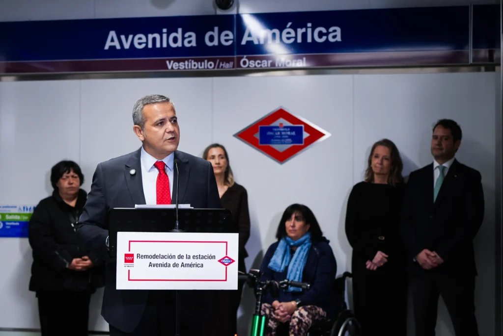 Un hombre trajeado habla en un podio rotulado Remodelación de la estación Avenida de América en el Metro de Avenida América, discutiendo la reforma para hacer la estación más accesible. Varias personas de pie y sentadas detrás de él, con un cartel de la estación encima.