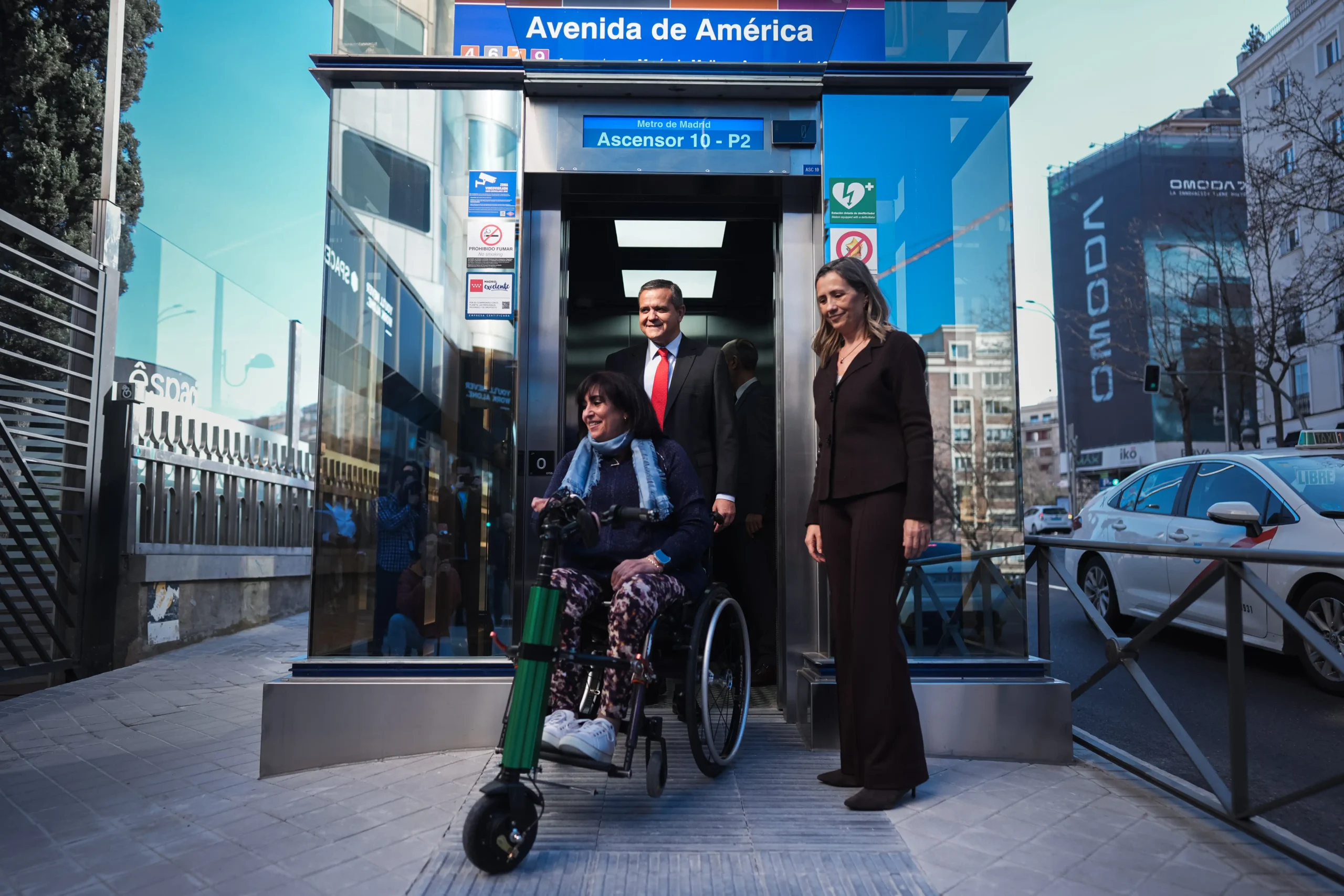 Una mujer en silla de ruedas sale de un ascensor accesible en la boca de Metro de Avenida América, acompañada por un hombre trajeado y una mujer vestida de oscuro traje de negocios en una calle de la ciudad.