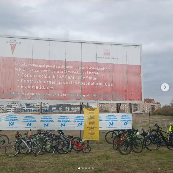 Un grupo de bicicletas aparca sobre el césped delante de una gran valla desgastada con texto en español sobre un proyecto de centro de salud en Rivas Vaciamadrid. Al fondo se ven edificios residenciales y un cielo nublado.