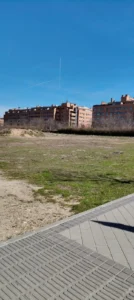 Una hilera de edificios de apartamentos marrones se alza tras un descampado cubierto de hierba y parcialmente sucio, con una acera asfaltada y una valla en primer plano y un cielo azul despejado en lo alto.