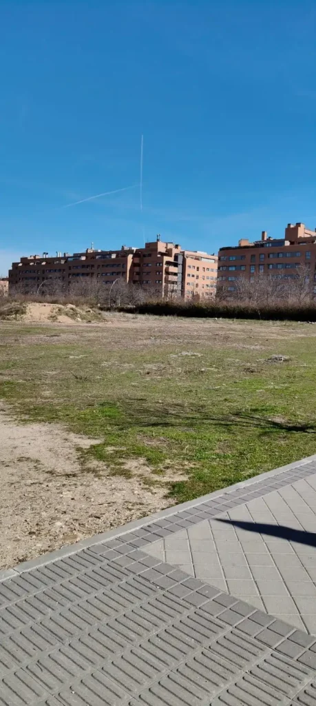 Una hilera de edificios de apartamentos marrones se alza tras un descampado cubierto de hierba y parcialmente sucio, con una acera asfaltada y una valla en primer plano y un cielo azul despejado en lo alto.
