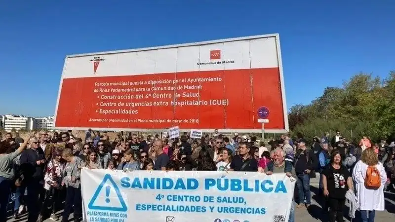Un grupo numeroso de personas frente a una valla sostiene una pancarta en la que se lee "Sanidad Pública, 4º Centro de Salud, Especialidades y Urgencias." La valla publicitaria de detrás hace referencia a la construcción de un cuarto centro de salud.