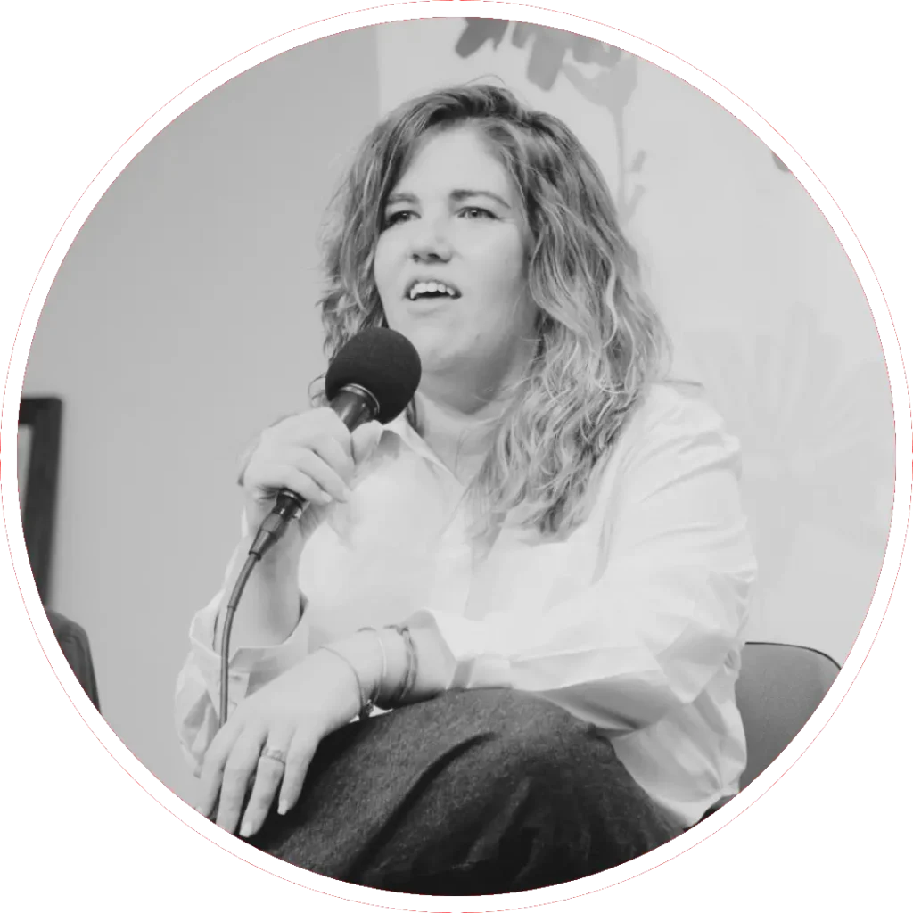 Yolanda Hidalgo es una mujer con el pelo ondulado está sentada sosteniendo un micrófono, lleva una camisa de color claro y pantalones oscuros. Parece estar hablando o respondiendo a preguntas en un debate. La imagen es en blanco y negro.