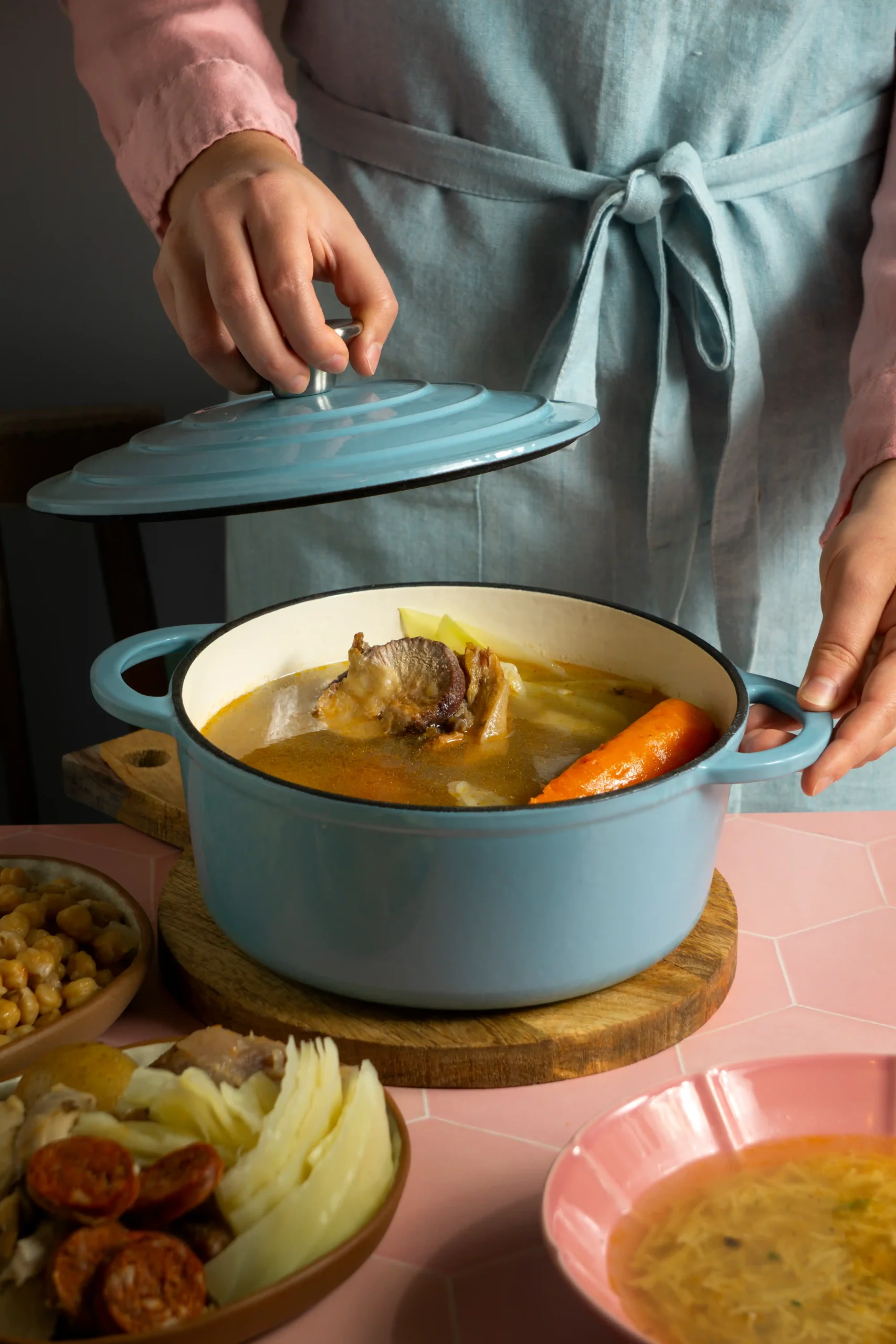 Una persona con un delantal azul claro levanta la tapa de una olla azul con sopa, dejando ver carne, una zanahoria y apio. Inspirada en la Ruta del Cocido 2026, la escena evoca sabores tradicionales celebrados en restaurantes de toda España. Una persona con un delantal azul claro levanta la tapa de una olla azul con sopa, dejando ver carne, una zanahoria y apio. Inspirada en la Ruta del Cocido 2026, la escena evoca sabores tradicionales celebrados en restaurantes de toda España.