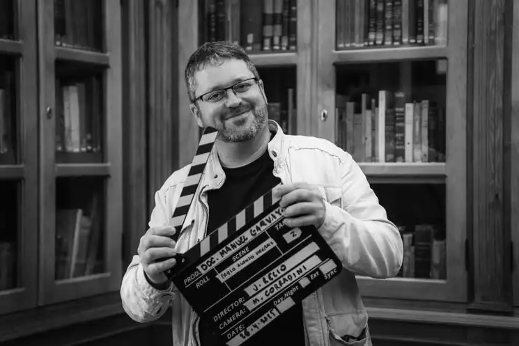 Un hombre con gafas y chaqueta clara sonríe mientras sostiene una claqueta de cine frente a una estantería llena de libros. La foto en blanco y negro evoca el espíritu de documentales sobre la Transición como "El hilo negro.