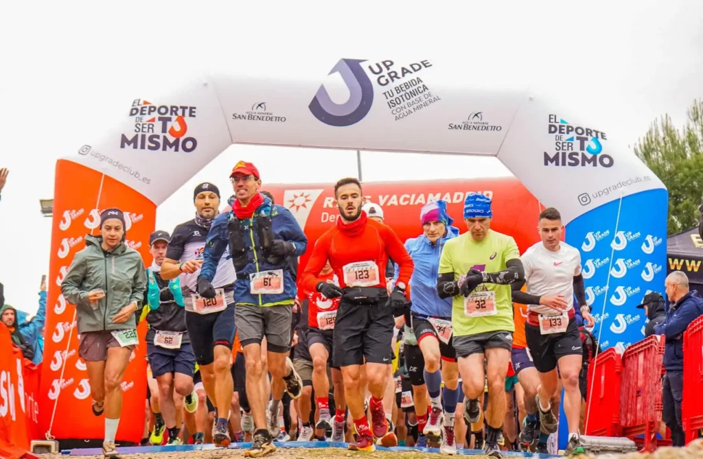 Corredores ataviados con ropa deportiva participan en la carrera Rivas Trail Natura, comenzando su recorrido bajo un arco hinchable en el que se exhiben los patrocinadores del evento y lemas escritos en español.
