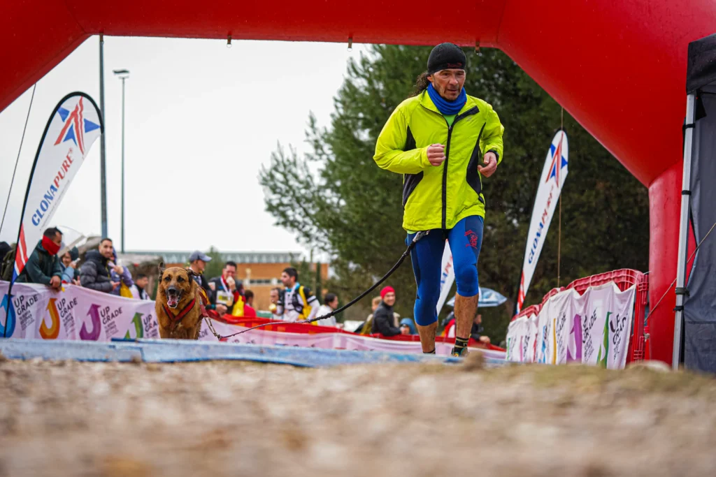 Un hombre con una chaqueta amarilla brillante y mallas azules corre con su perro marrón bajo un arco hinchable rojo durante la carrera Rivas Trail Natura, acompañado por otros corredores, mientras se ven espectadores y pancartas del evento al fondo.