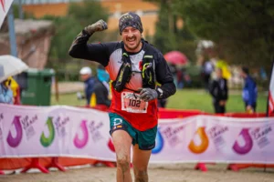 Un corredor con barba y bigote, con el dorsal 102, gorra negra y ropa deportiva embarrada, sonríe y levanta el puño mientras cruza la meta del Rivas Trail Natura bajo la lluvia con otros corredores en un día lluvioso.