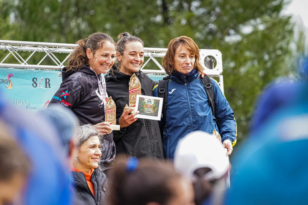 Cuatro mujeres suben a un podio al aire libre en el Rivas Trail Natura, tres sostienen trofeos de madera y una un certificado enmarcado. Las corredoras van vestidas con ropa deportiva, con árboles y una pancarta promocional de fondo.