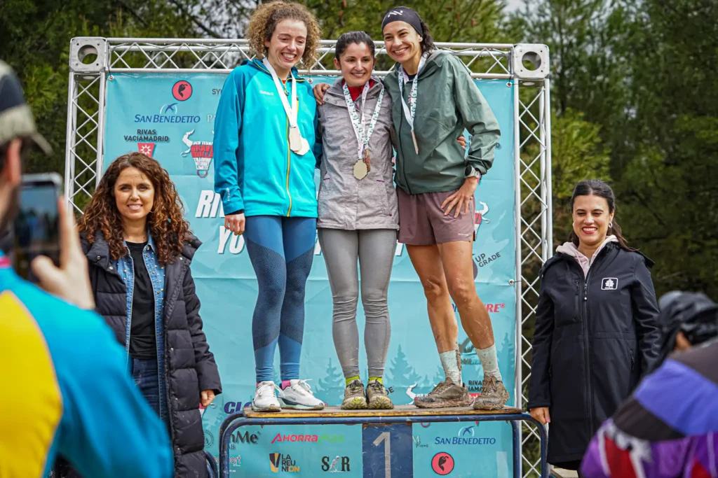 Tres mujeres corredoras con medallas suben a un podio para la entrega de premios de la carrera Rivas Trail Natura, con una mujer en el centro en la posición más alta. Dos mujeres están a cada lado mientras alguien delante capta el momento con una foto.