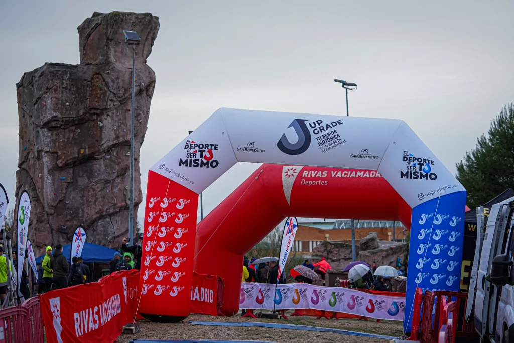 Arco hinchable en un evento al aire libre de Rivas Trail Natura, con logotipos y texto. Corredores con chaquetas y sombreros se reúnen cerca del arco, que está rodeado de vallas. Al fondo, una gran formación rocosa, a pesar de la amenaza de lluvia.
