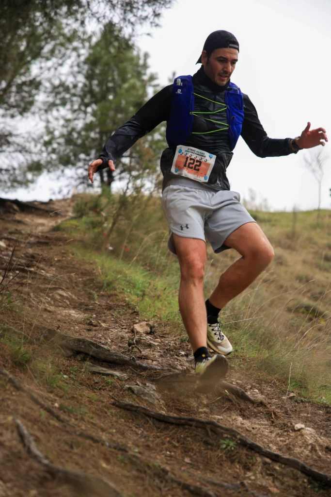 Un hombre con dorsal, chaleco de corredor, camiseta negra de manga larga, pantalón corto gris y zapatillas de trail desciende por un empinado e irregular camino de tierra durante la carrera Rivas Trail Natura. Al fondo se ven otros corredores y frondosos árboles y hierba.
