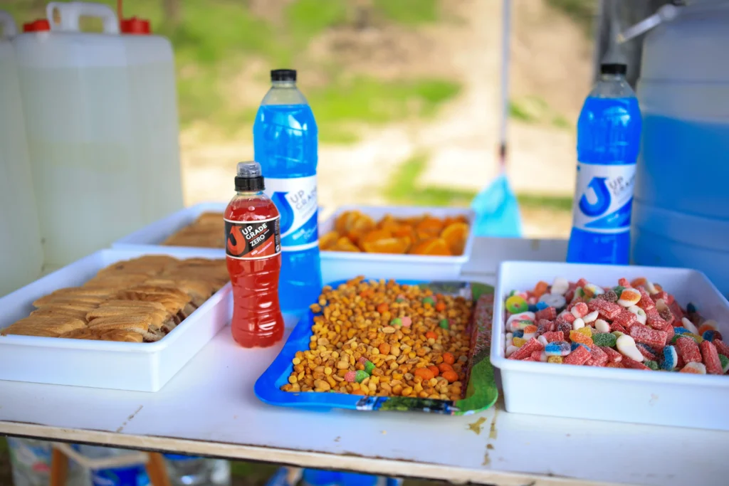 Una mesa muestra bandejas de bollería, frutos secos variados, gominolas confitadas y rodajas de naranja para los corredores de Rivas Trail Natura, junto con botellas de bebidas azules y rojas. Al fondo se ven grandes contenedores de agua a pesar de la lluvia.