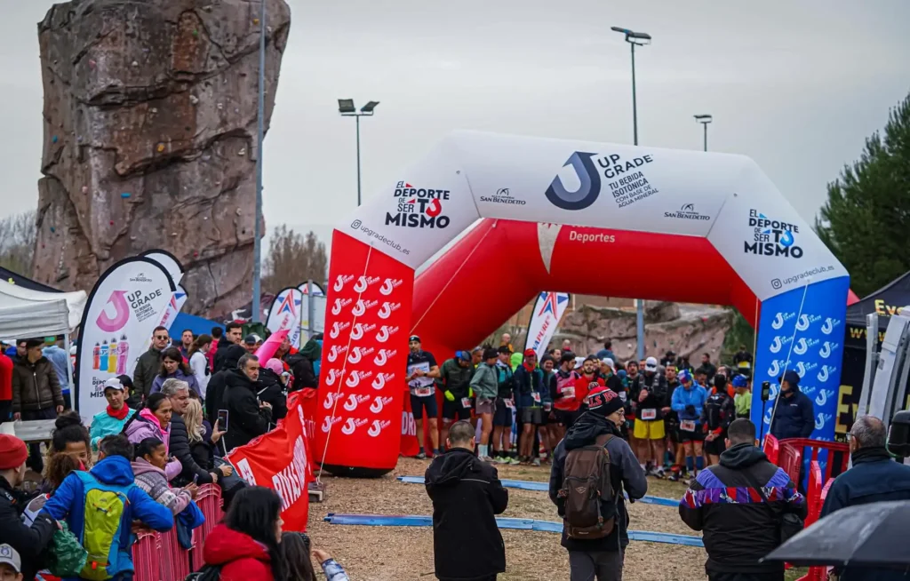 Una multitud se reúne en torno a un grupo de corredores en la línea de salida del Rivas Trail Natura, marcada por un arco hinchable blanco y rojo con varias pancartas y banderas. Algunos espectadores hacen fotos mientras otros observan.