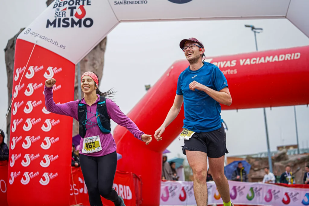 Dos corredores, una mujer y un hombre, se cogen de la mano y sonríen mientras cruzan la línea de meta roja y blanca del Rivas Trail Natura con atuendo deportivo y dorsal. La mujer levanta el puño en señal de celebración a pesar de la ligera lluvia.