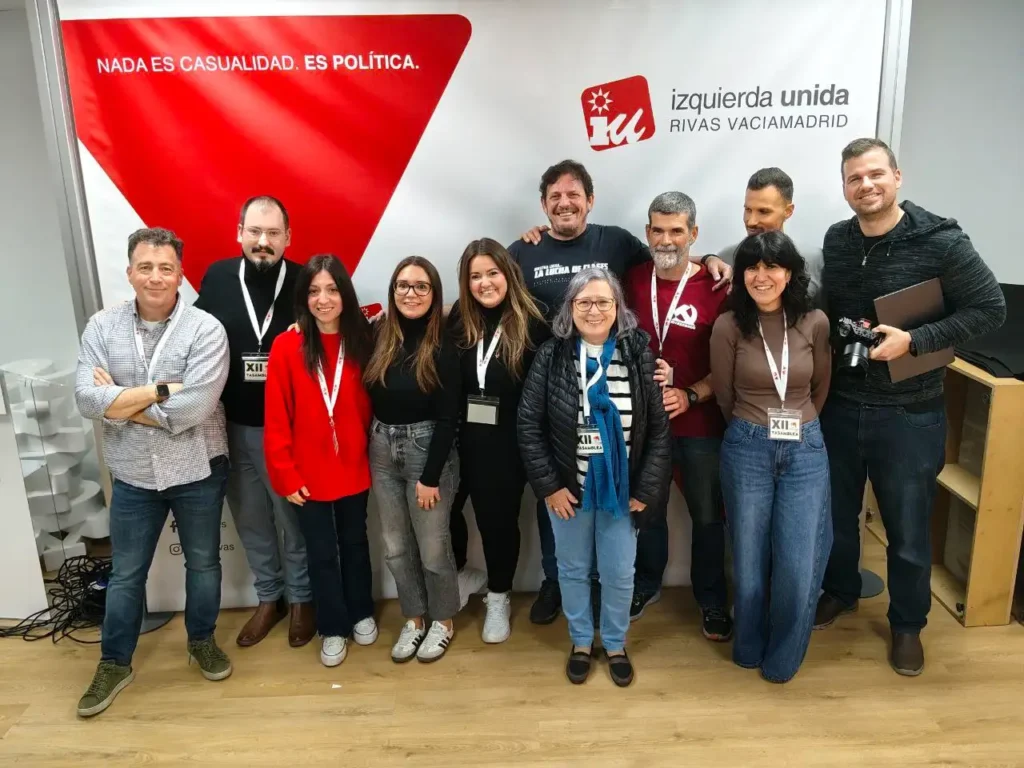 Un grupo de doce personas, algunas con cordones y chapas, posan para una foto delante de una pancarta en la que se lee IU Rivas Vaciamadrid. La coordinadora, Aída Castillejo, se sitúa entre ellos junto al triángulo rojo y el texto en español.
