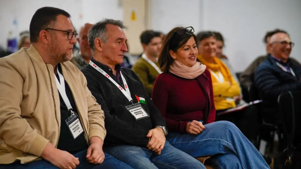 Un grupo de personas con distintivos de la conferencia se sientan en sillas, mirando al frente y escuchando atentamente. La atención se centra en la coordinadora Aída Castillejo y otras personas de IU Rivas en primera fila, con más asistentes sentados detrás de ellas en un luminoso ambiente interior.
