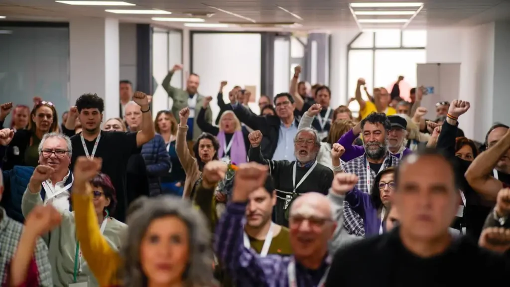 Un nutrido grupo de personas en el interior, muchas de ellas levantando el puño en alto, permanecen juntas en un pasillo o sala de conferencias muy iluminada, mostrando su apoyo a la coordinadora Aída Castillejo y a IU Rivas.