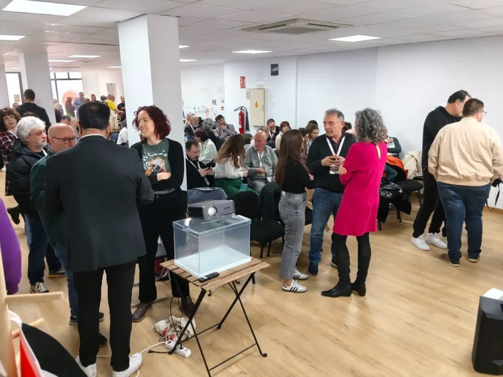 Un grupo de personas está reunido en una habitación luminosa con paredes blancas y suelos de madera. Algunas están de pie y hablando, mientras que otras están sentadas. Sobre una mesa hay una urna transparente y la coordinadora Aída Castillejo, de IU Rivas, está presente.