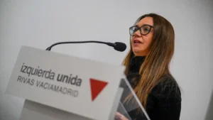 Aída Castillejo, coordinadora de IU Rivas, con pelo largo castaño y gafas, habla en un estrado rotulado izquierda unida RIVAS VACIAMADRID con un logotipo de un triángulo rojo, dirigiéndose al micrófono sobre un fondo liso y claro.