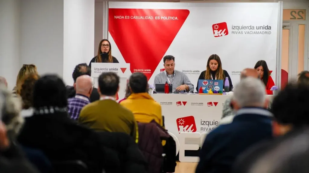 Aída Castillejo, coordinadora de IU Rivas, habla en un podio a un público en una sala de conferencias, con tres personas sentadas en una mesa a su lado. Por toda la sala se ven pancartas y carteles con "izquierda unida" y gráficos rojos.