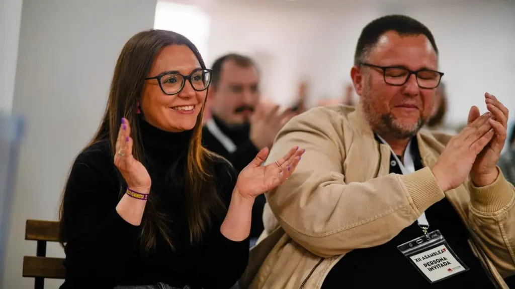 Dos personas aplauden sentadas una al lado de la otra, ambas con gafas. La mujer de la izquierda, Aída Castillejo, coordinadora de IU Rivas, sonríe con un jersey negro de cuello alto, mientras que el hombre de la derecha lleva una chaqueta beige y una placa con su nombre. Otros aparecen difuminados en el fondo.